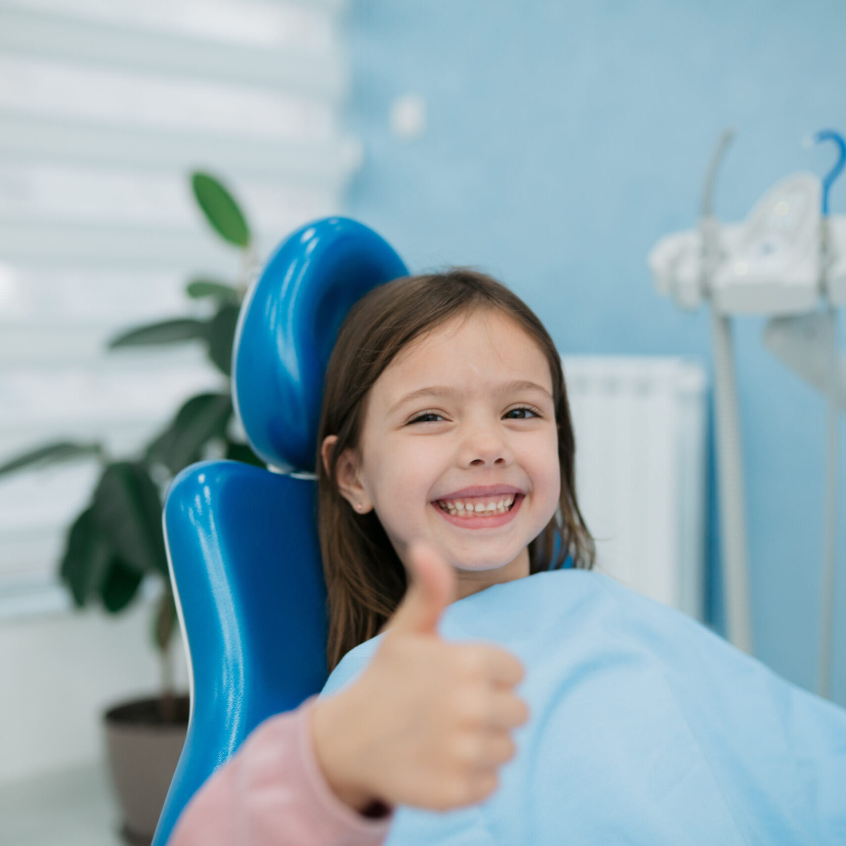 Portrait of a little happy girl sitting in a dentist chair with a toothy smile on her face, looking at the camera and showing a thumbs up gesture with her hand